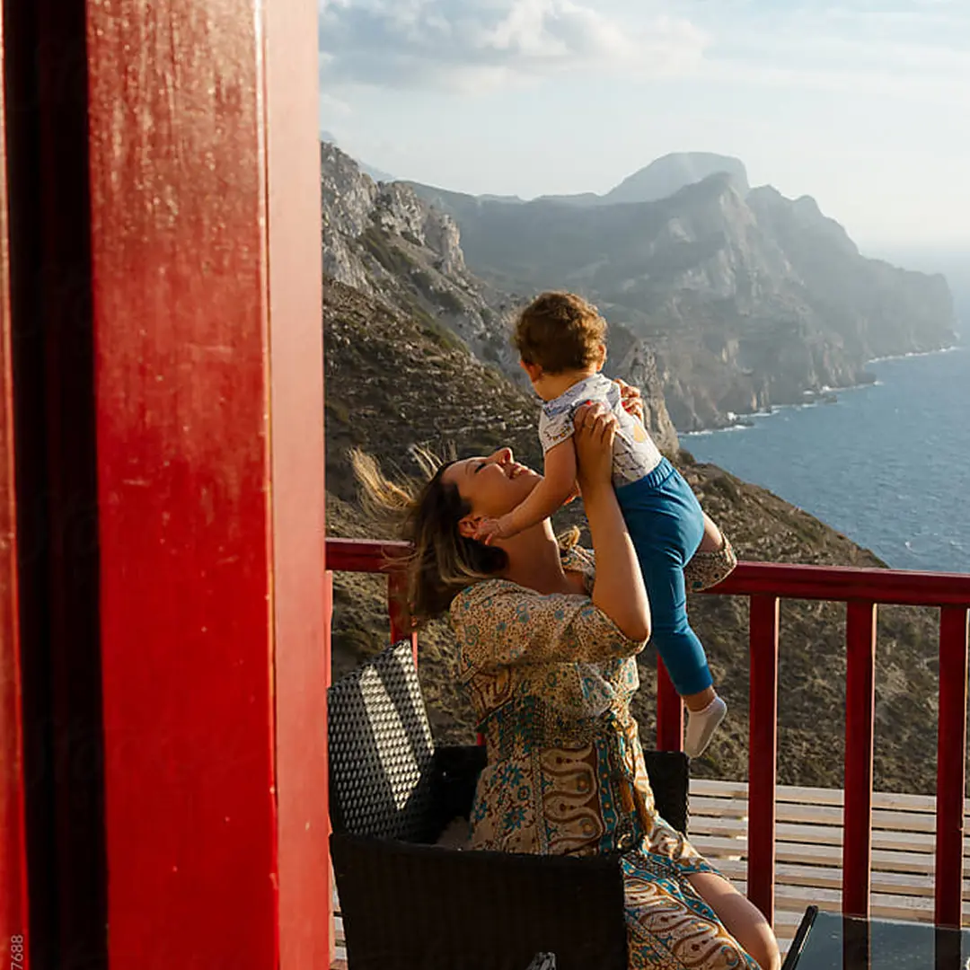 Woman joyfully lifts a child on a sunlit balcony overlooking a scenic coastal cliff and ocean under a blue sky with clouds.