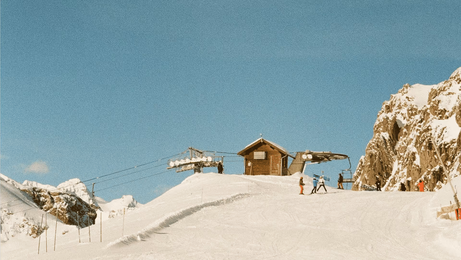 Skiers on a snowy slope approach a wooden ski lift station under a clear blue sky. Rocky, snow-covered peaks surround the serene alpine scene.
