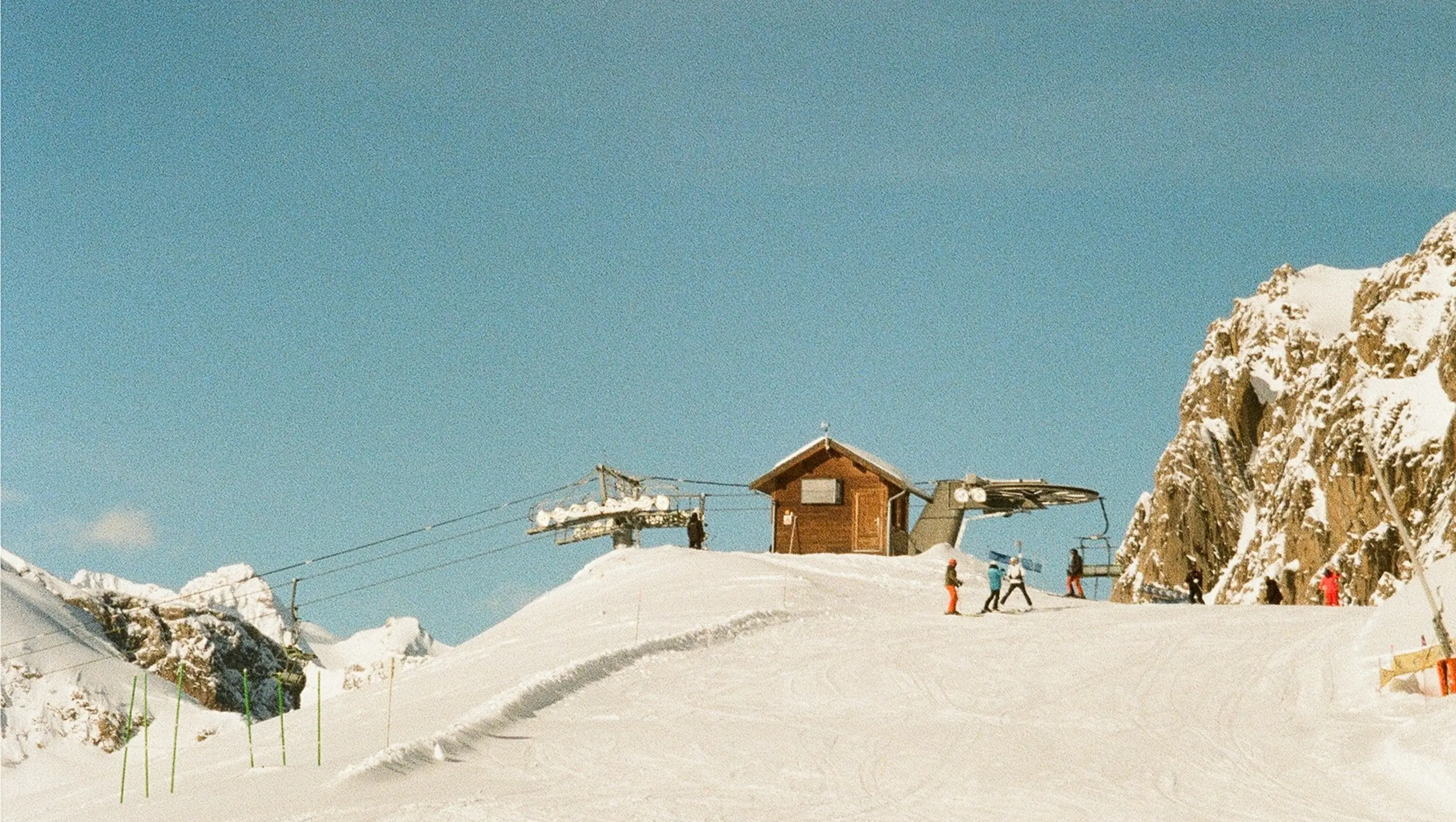 Skiers on a snowy slope approach a wooden ski lift station under a clear blue sky. Rocky, snow-covered peaks surround the serene alpine scene.