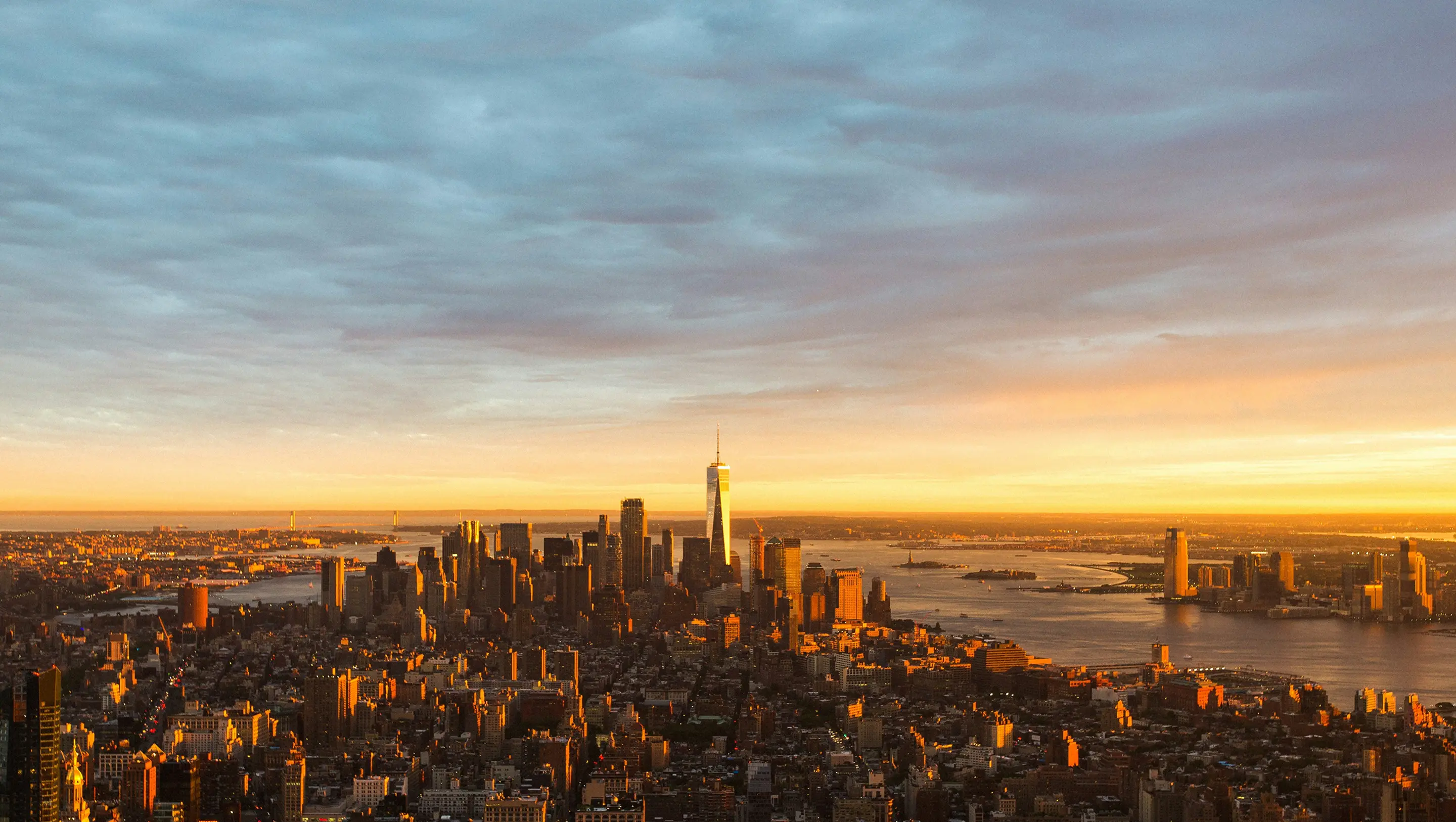 Aerial view of downtown Manhattan during sunset, with skyscrapers illuminated by golden light. The Hudson River reflects the warm hues, while clouds fill the sky.