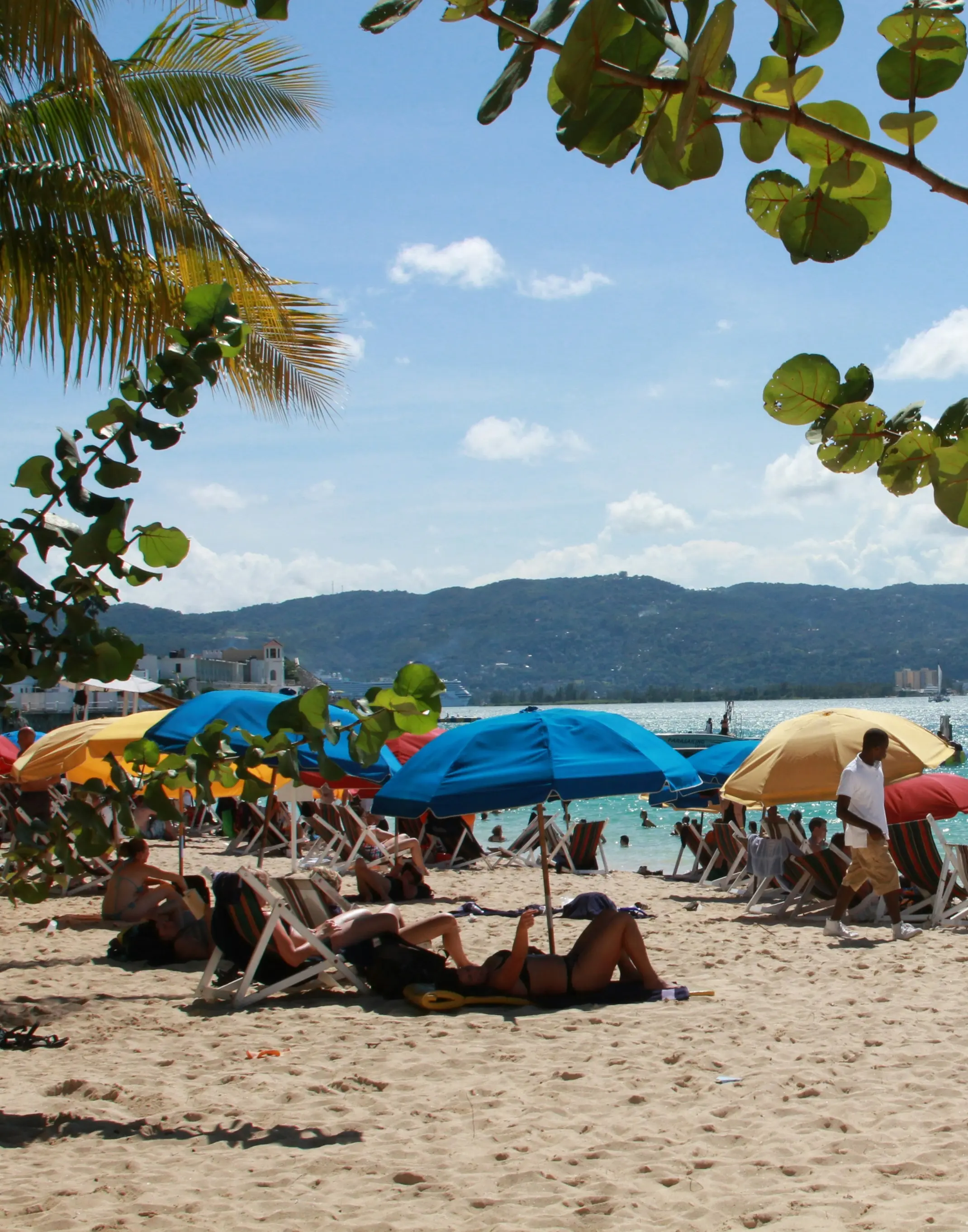 A sunny Montego Bay beach scene with people relaxing under colorful umbrellas. Palm leaves frame a view of turquoise water and mountains, creating a tropical vibe.