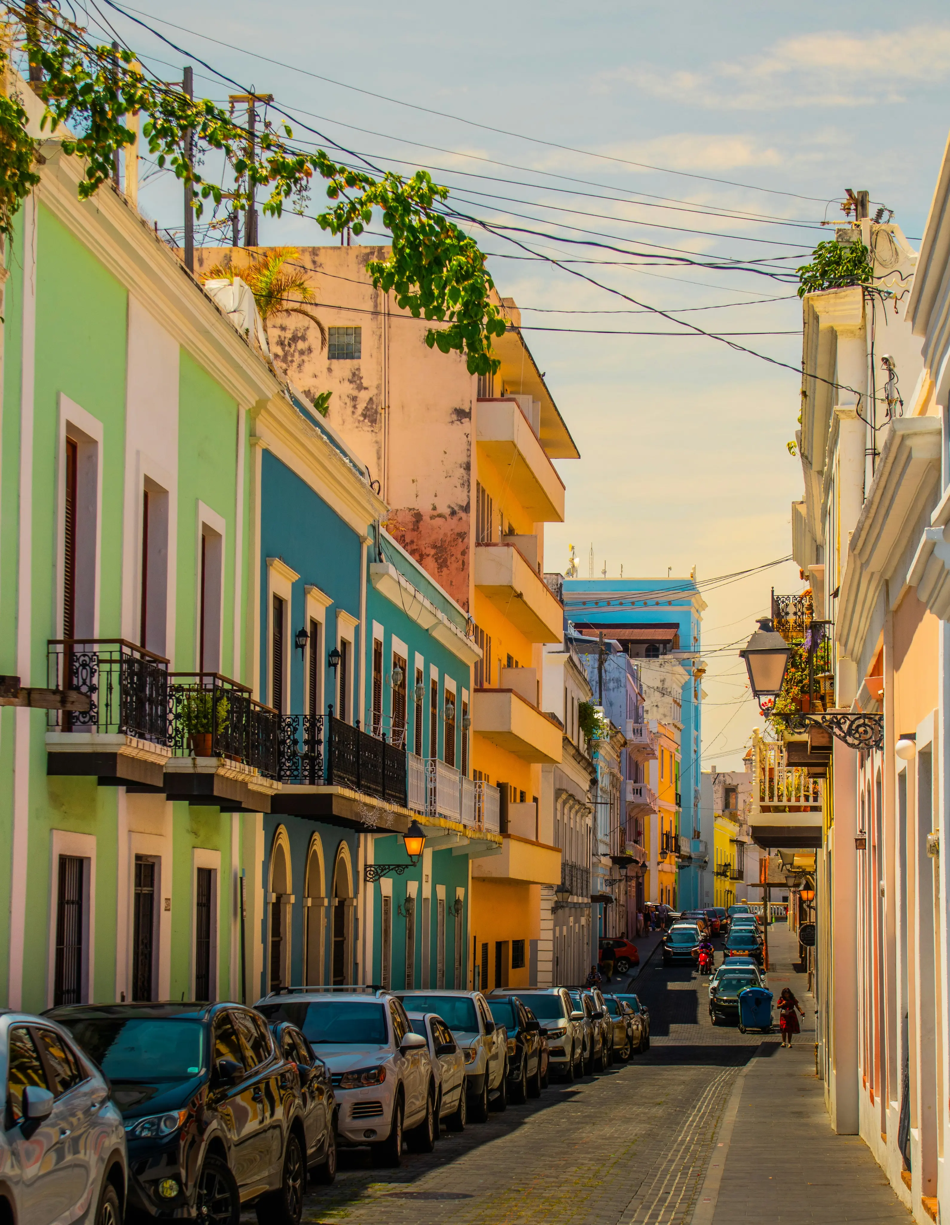 A picturesque street in Old San Juan, with colorful buildings in pastel shades of green, blue, and yellow. Cars line one side, while the sunny sky adds warmth.