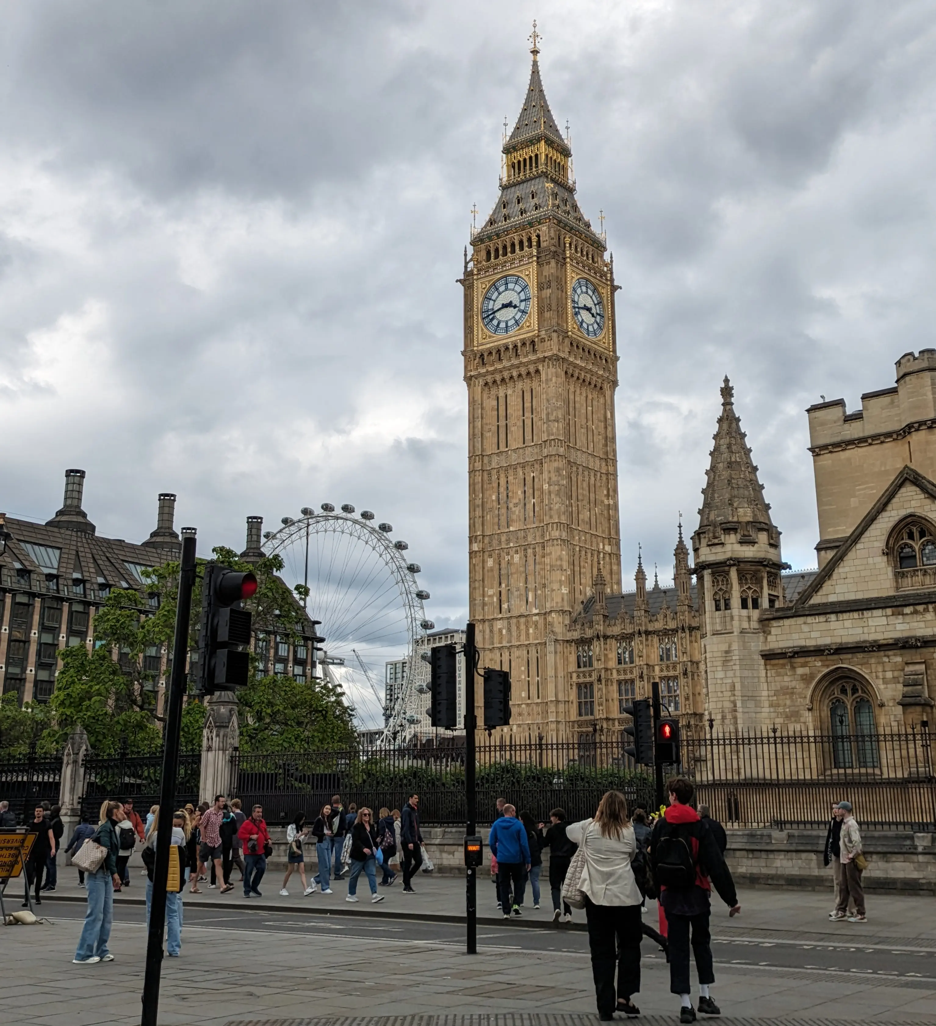 Big Ben and the London Eye under a cloudy sky, with people walking and waiting at a crosswalk. The vibe is busy and iconic London.