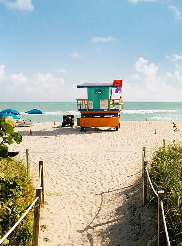 Pathway leading to a sandy beach with a colorful lifeguard hut. Blue ocean waves, umbrellas, and sunny skies create a vibrant, relaxed atmosphere.