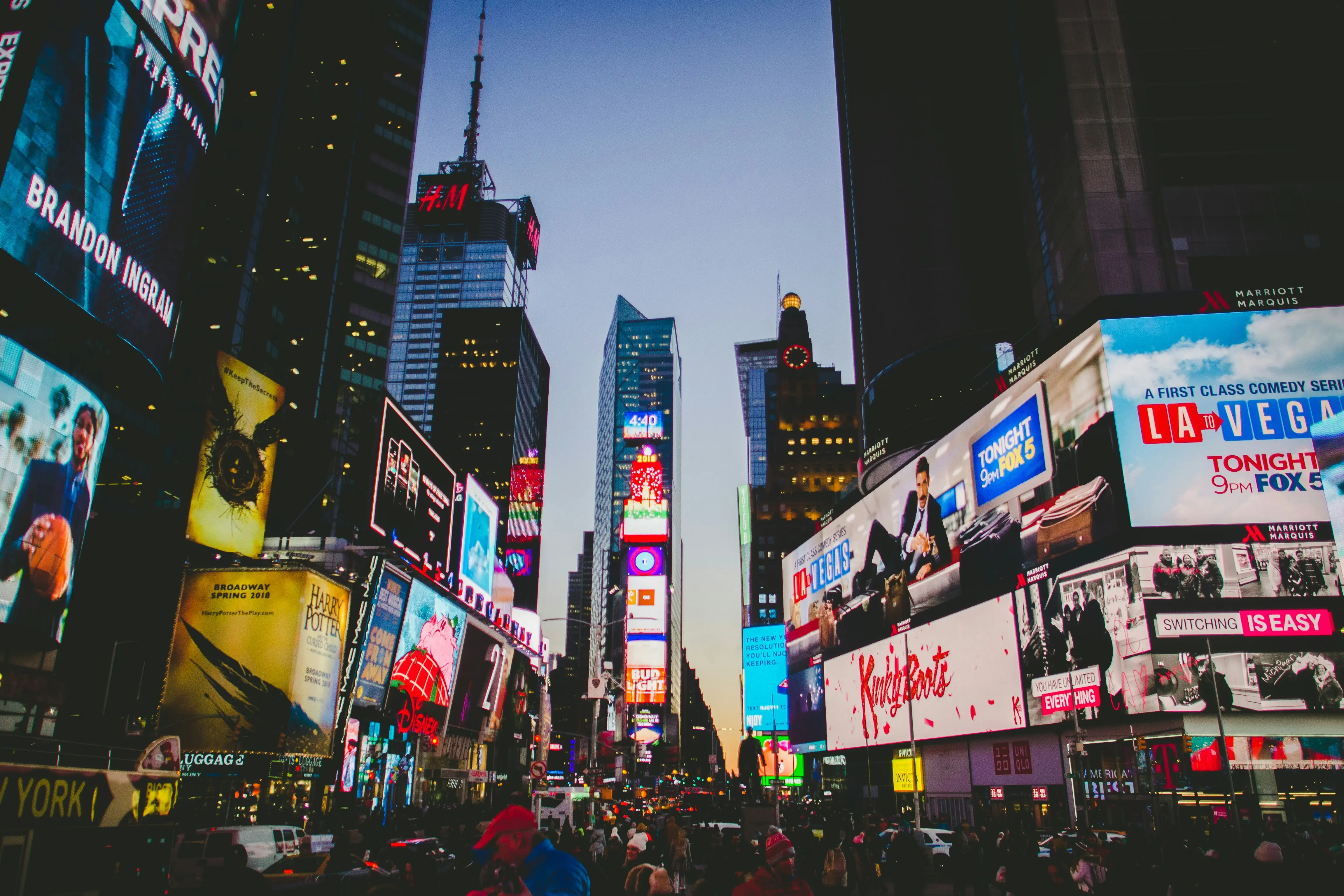 Bright, colorful billboards light up Times Square at dusk, featuring advertisements and neon lights. The bustling street below teems with people and vehicles.