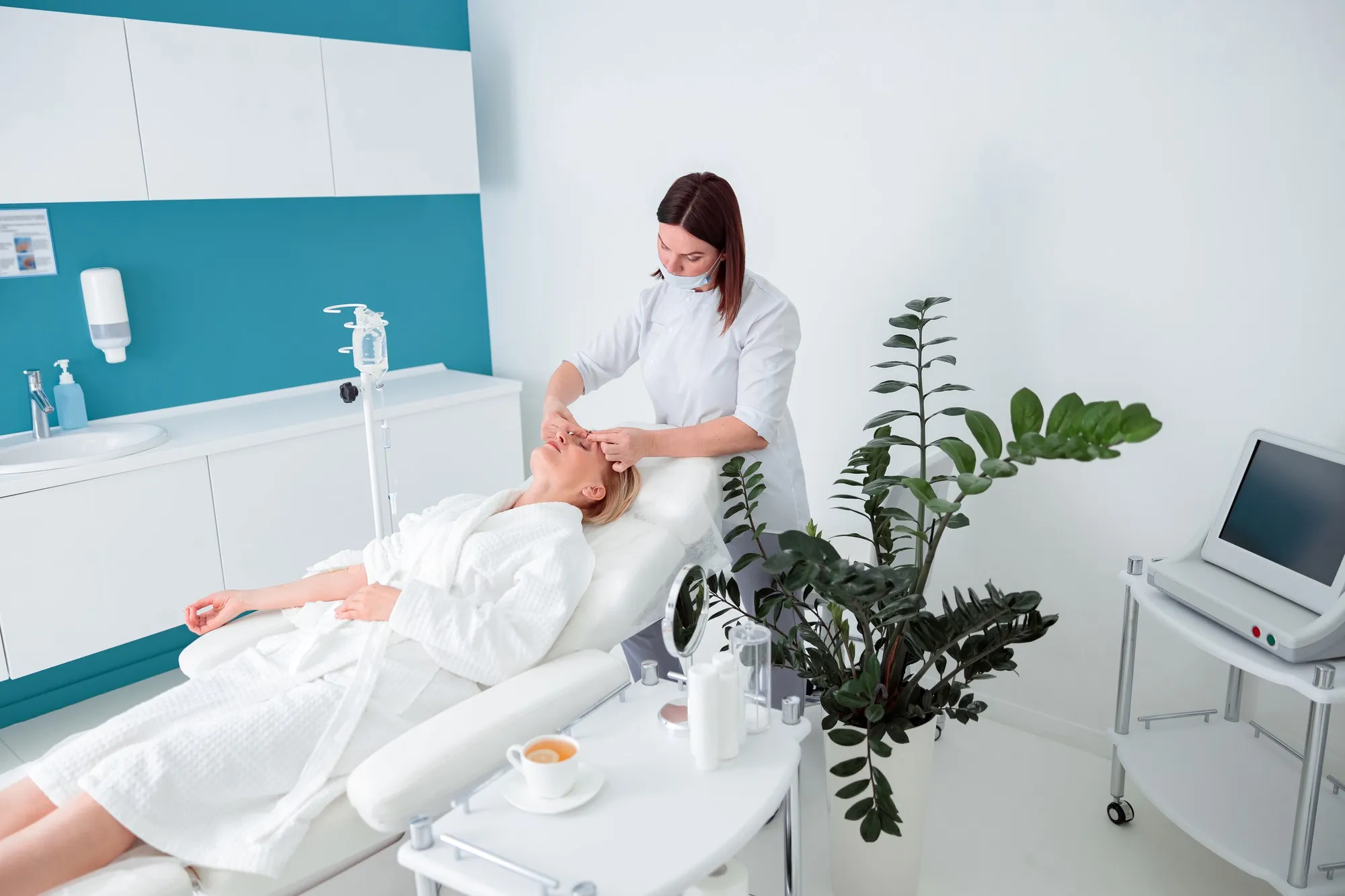 Kvinne får ansiktsbehandling av en hudpleier i en moderne klinikk. Hun ligger avslappet i en behandlingsstol iført hvit morgenkåpe, omgitt av medisinsk utstyr, planter og et bord med speil og produkter.
