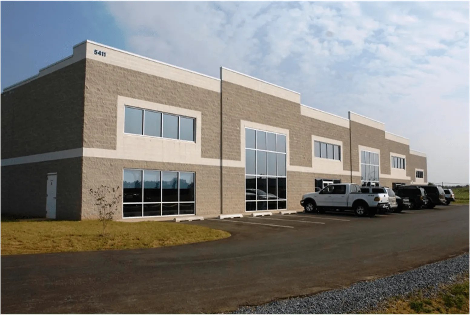 Modern commercial building with large glass windows and parked vehicles along the driveway.
