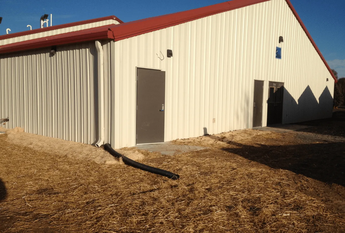 Exterior view of a metal storage building with a red roof and two doors, surrounded by straw-covered ground.
