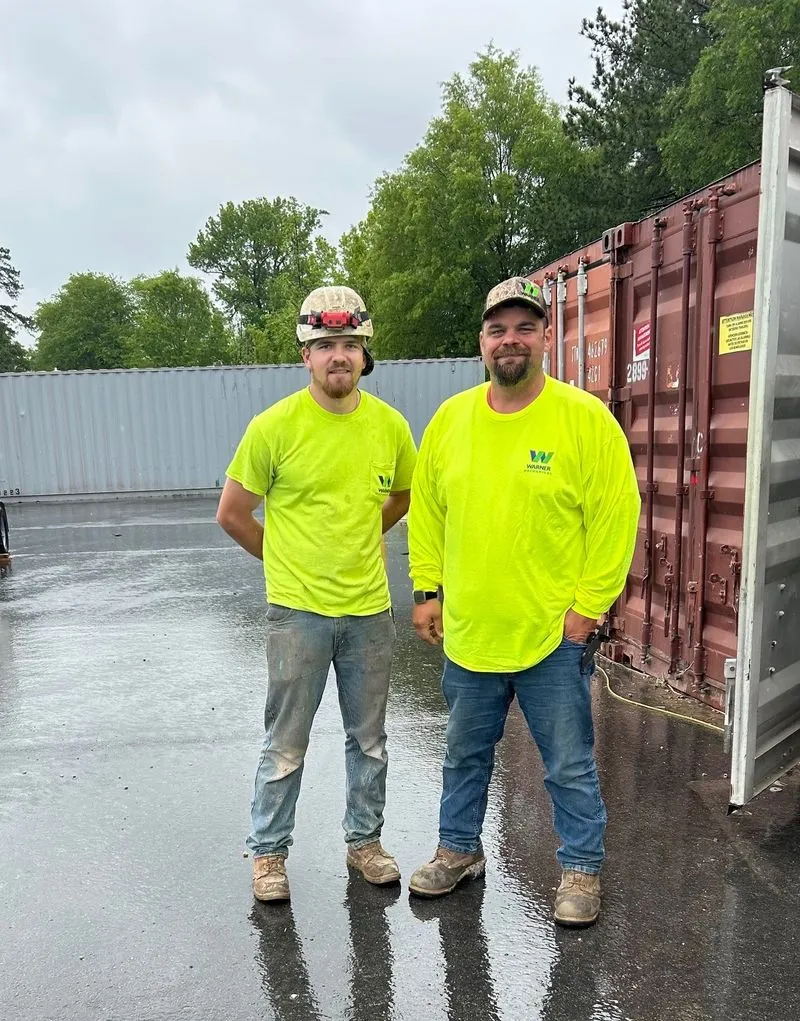 Two construction workers in bright yellow shirts stand outdoors near storage containers on a rainy day.
