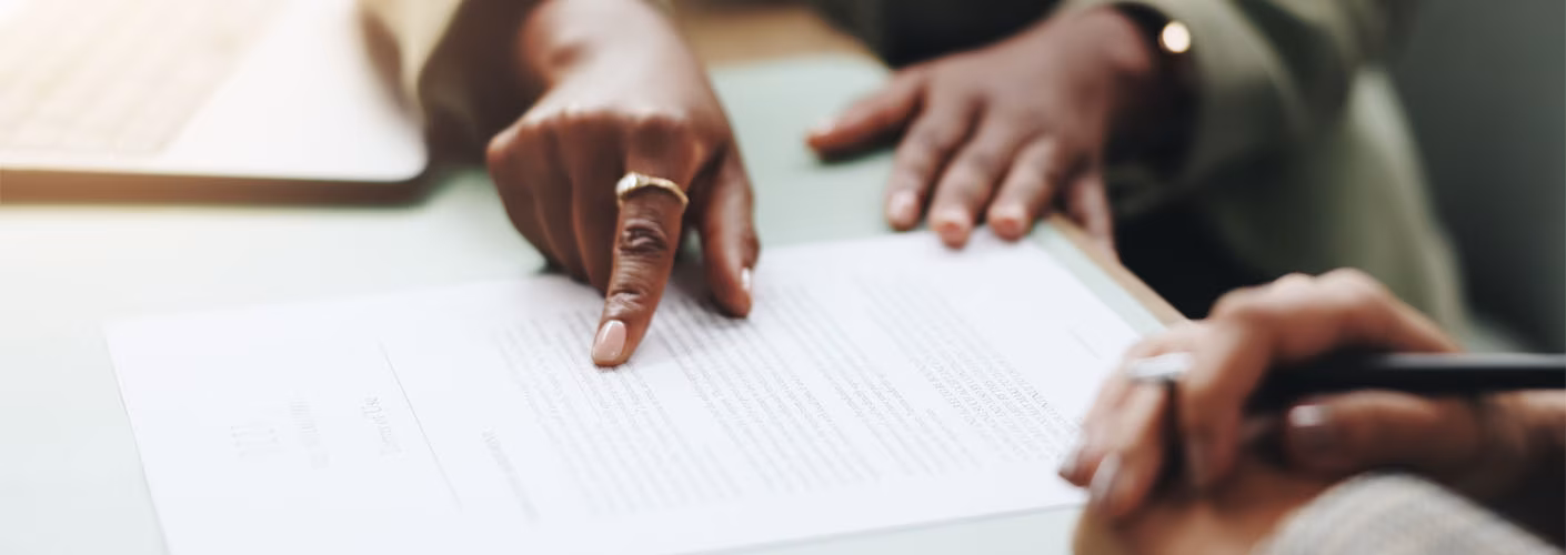 A hand pointing at a paper on a desk with text on it while another hand holds a pen