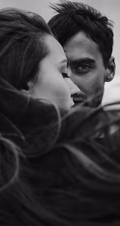 Young couple in black and white with man looking into camera with one eye while other part of his face is aesthetically covered with his ladies face and long hair observed from back-side.