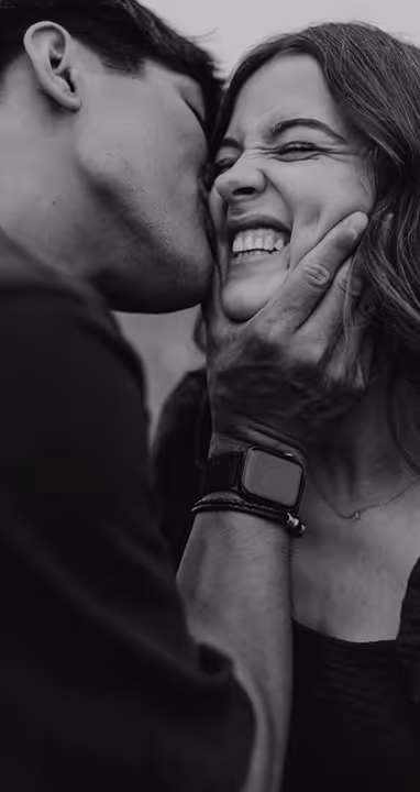 Young couple humorous with him as if grabbing her by chin and kissing into her cheek while her is laughing with closed eyes and smiling lips, her teeth visible due her smile.