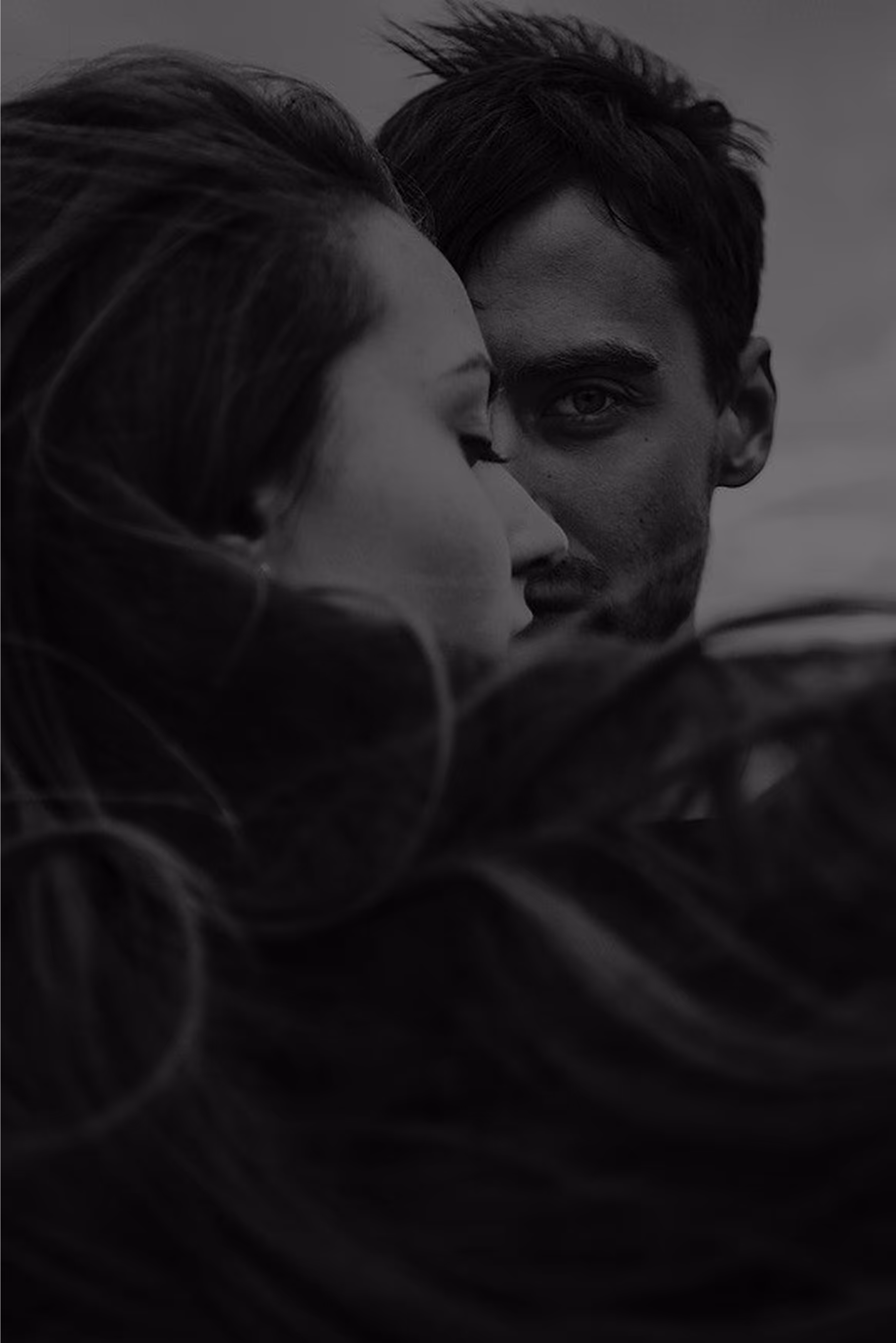 Young couple in black and white with man looking into camera with one eye while other part of his face is aesthetically covered with his ladies face and long hair observed from back-side.