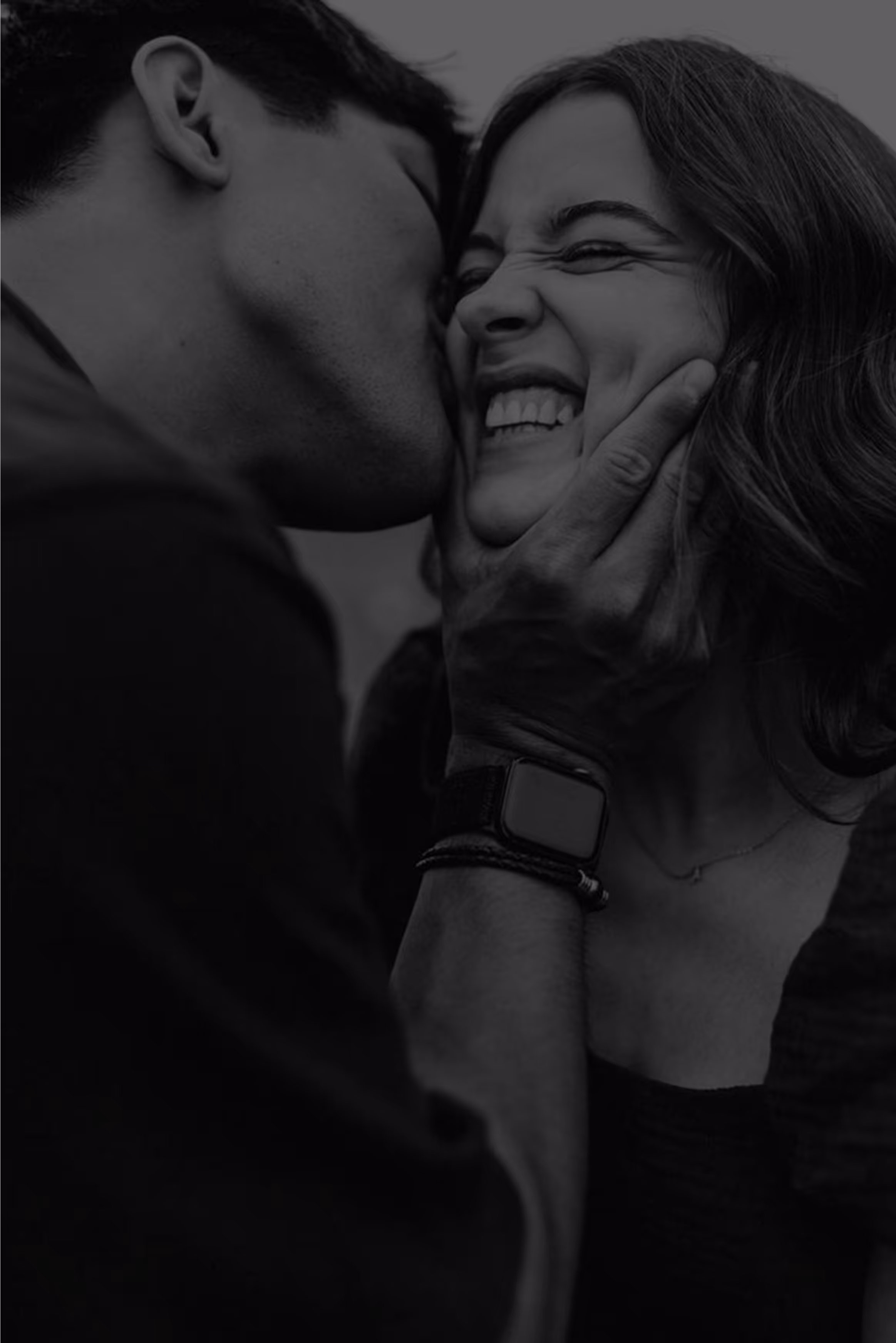 Young couple humorous with him as if grabbing her by chin and kissing into her cheek while her is laughing with closed eyes and smiling lips, her teeth visible due her smile.
