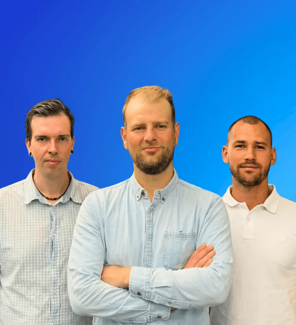 Drei Männer vor einem blauen Hintergrund, der Mann in der Mitte trägt ein hellblaues Hemd mit verschränkten Armen.