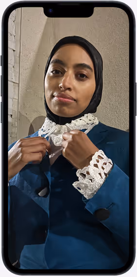 Porträt einer Frau mit schwarzem Kopftuch und blauem Anzugjacke mit weißen gehäkelten Spitzen am Kragen und Ärmelmanschetten, aufgenommen auf dem Bildschirm eines Smartphones.