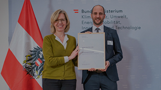 Eine Frau und ein Mann halten gemeinsam eine Urkunde und lächeln in dir Kamera.