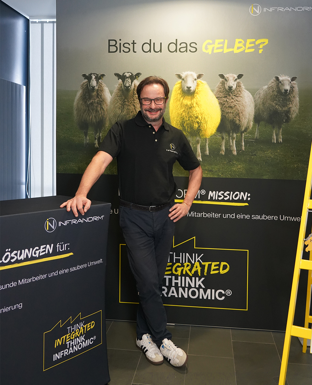 Christian Lindner, der Geschäftsführer von Infranorm, steht vor einem Plakat von Infranorm und stützt sich mit einer Hand an einem Tisch ab.