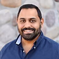 Smiling man with beard wearing a blue shirt standing in front of a stone wall.