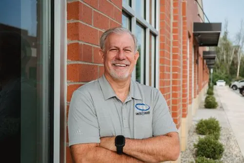 Smiling older man with gray hair and beard leaning against brick wall outside office building.