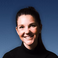 Smiling woman with brown hair pulled back wearing a black top against a blue background.