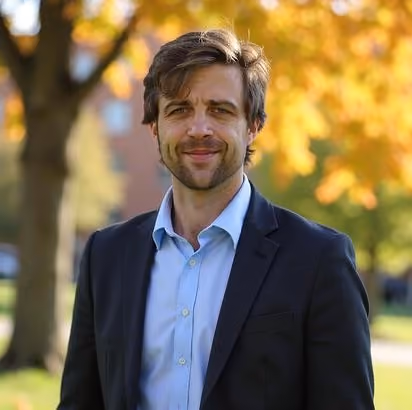 Smiling man in a black blazer and light blue shirt standing outdoors with autumn trees in the background.
