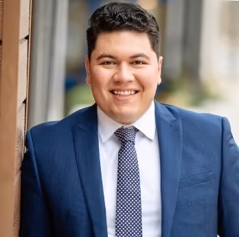 Smiling man in a blue suit and polka dot tie leaning against a wall outdoors.