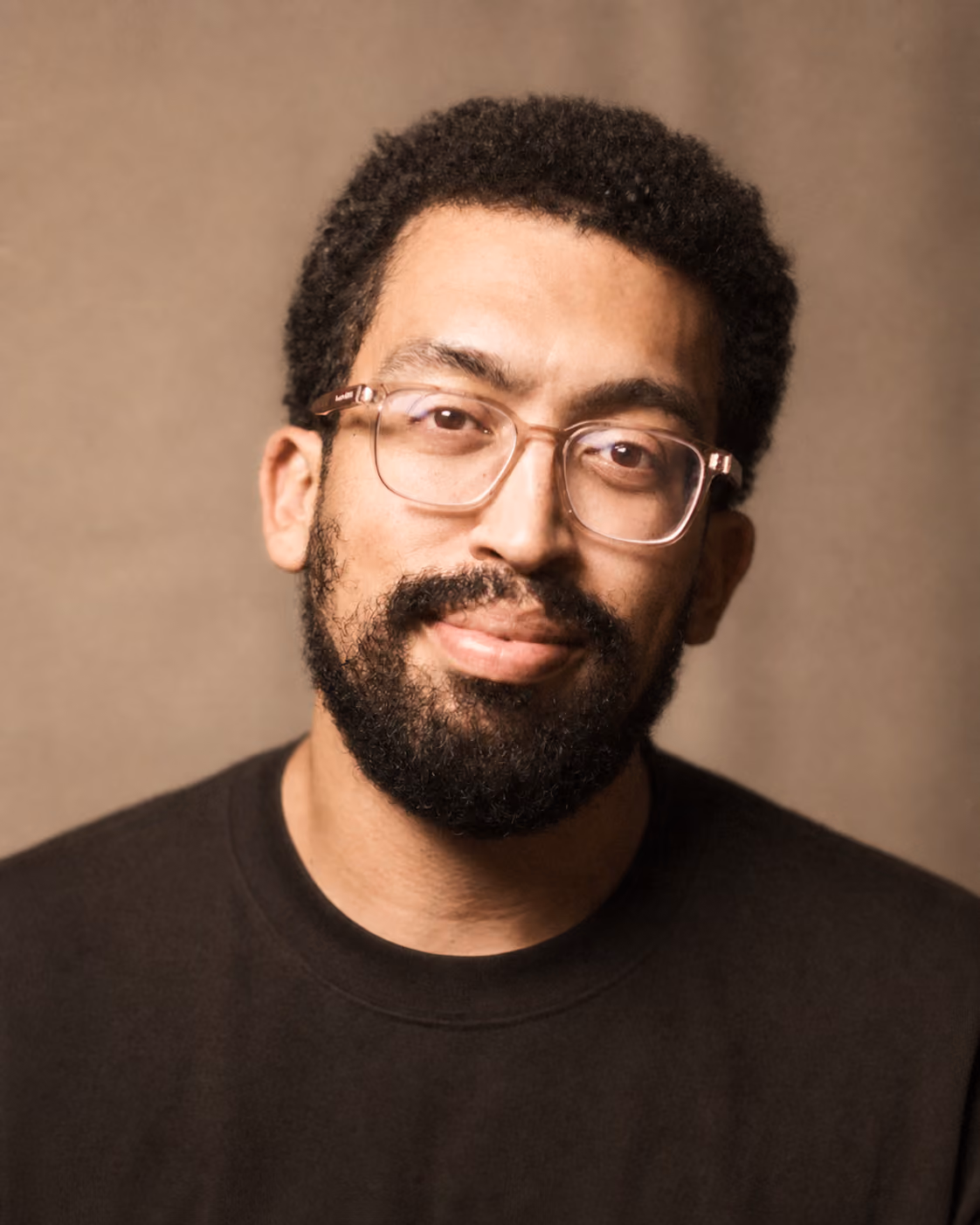 Smiling man with curly hair, glasses, and a beard wearing a black shirt against a neutral background.