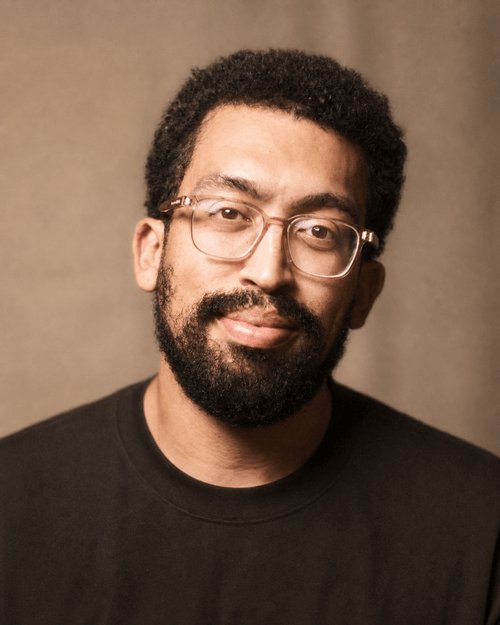 Smiling man with curly hair, glasses, and a beard wearing a black shirt against a neutral background.