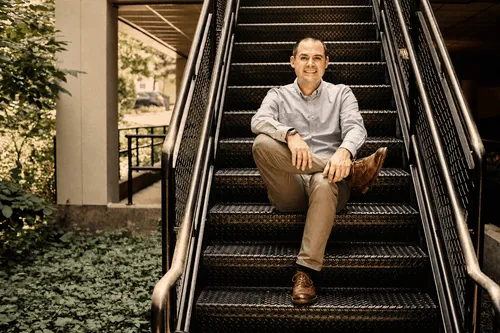 Man in light gray shirt and brown shoes sitting cross-legged on black metal stairs outdoors.