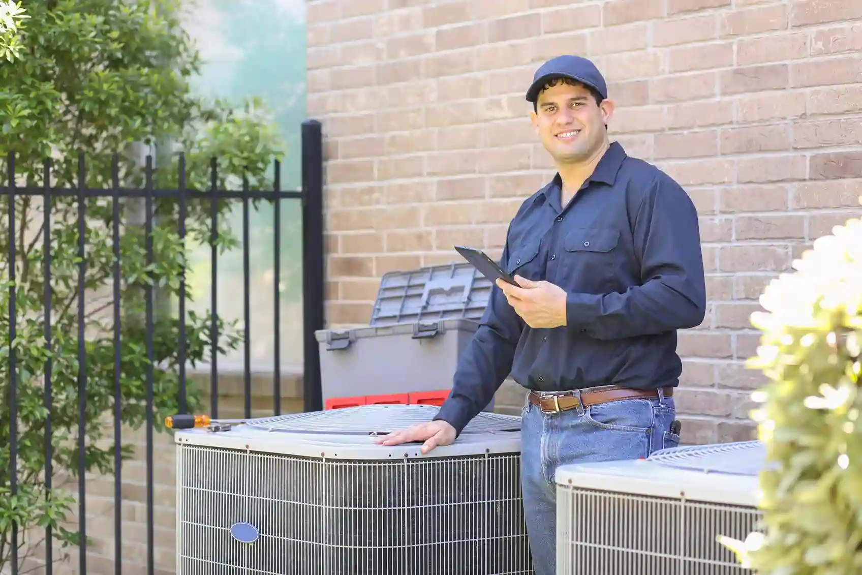 Technician in dark blue shirt using tablet beside two outdoor AC units, with toolbox on top, near brick wall and greenery