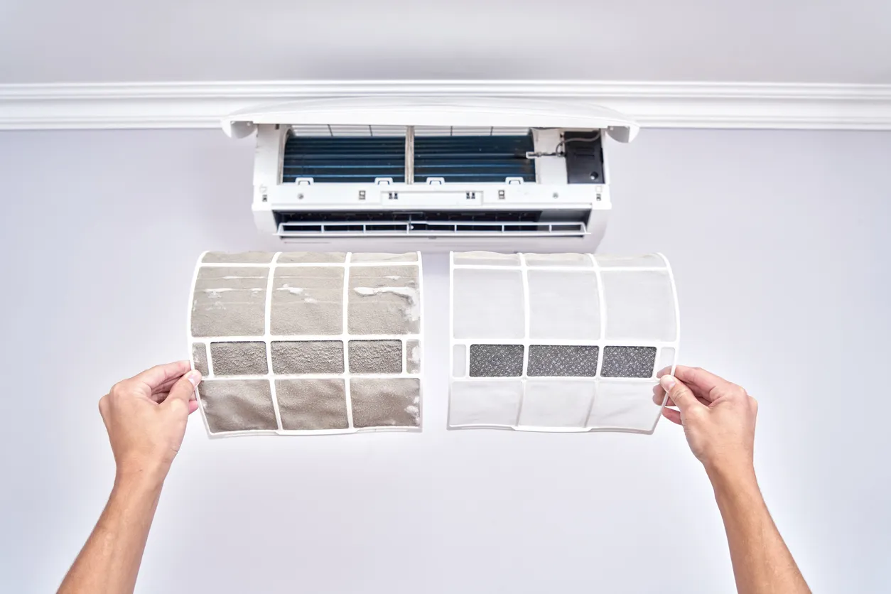 Person holding dirty and clean air filters in front of open wall-mounted air conditioner