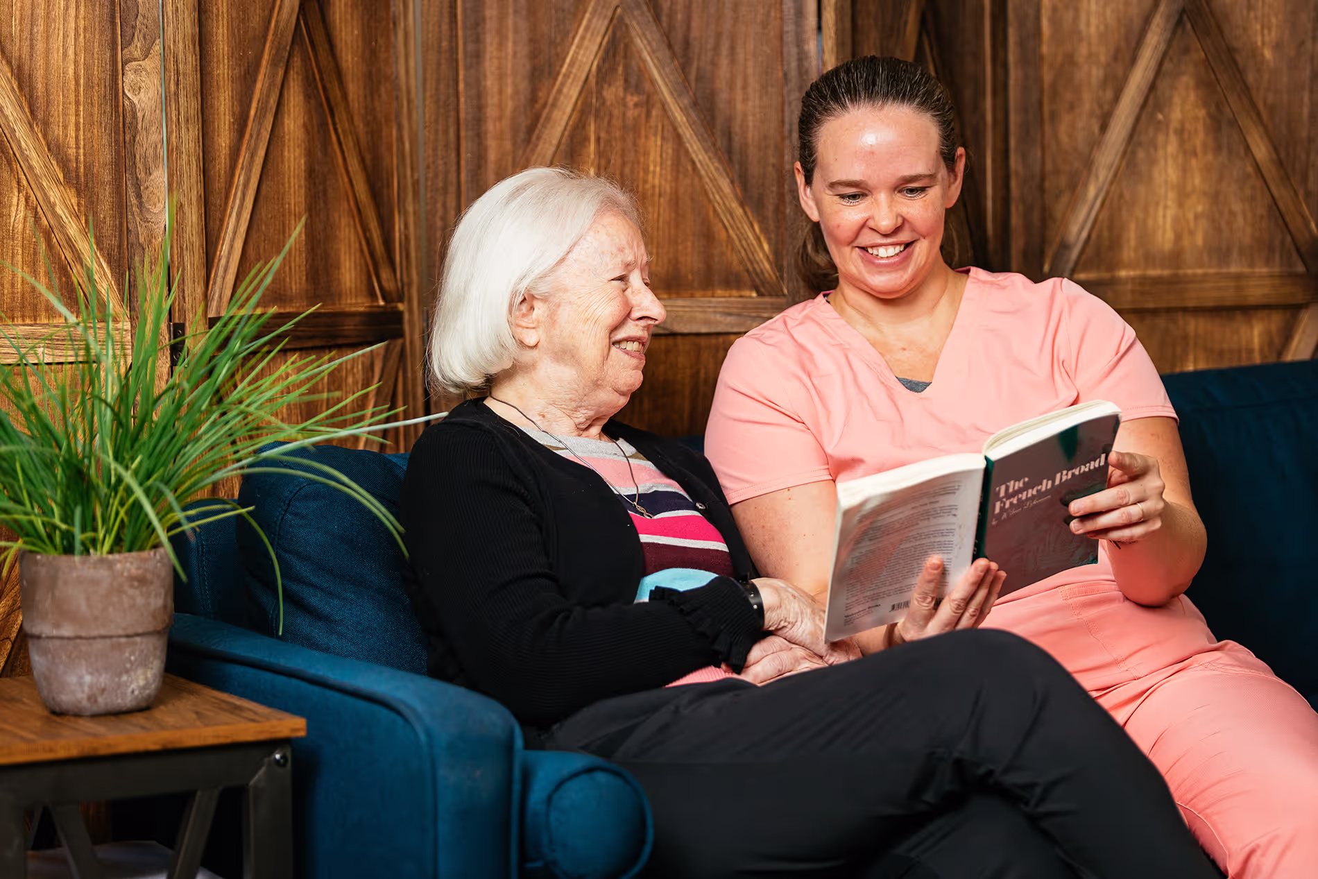 An older woman and a younger woman sit together on a couch, smiling and reading a book.