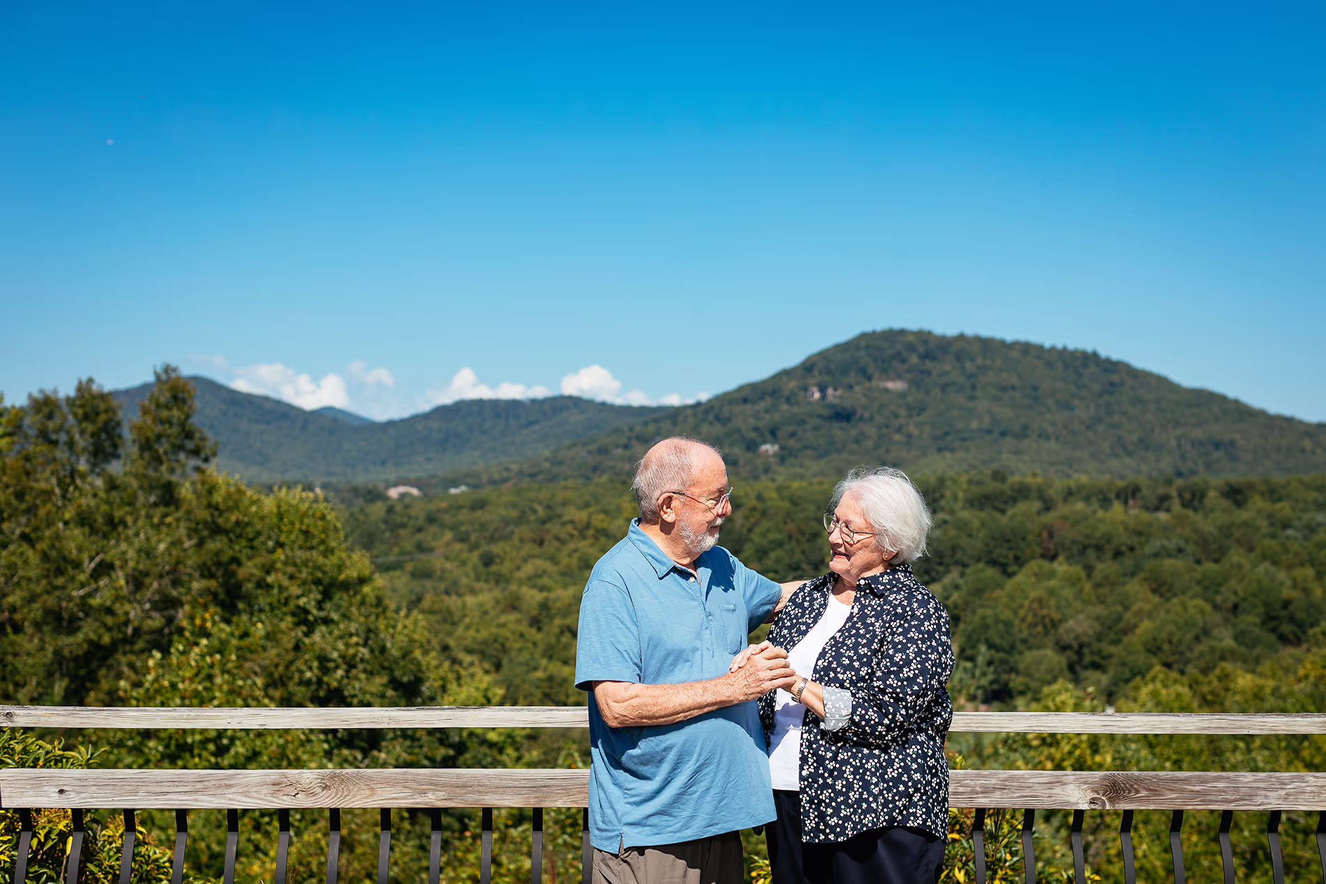 An elderly couple stands on a wooden deck, smiling and holding hands, with mountains in the background.