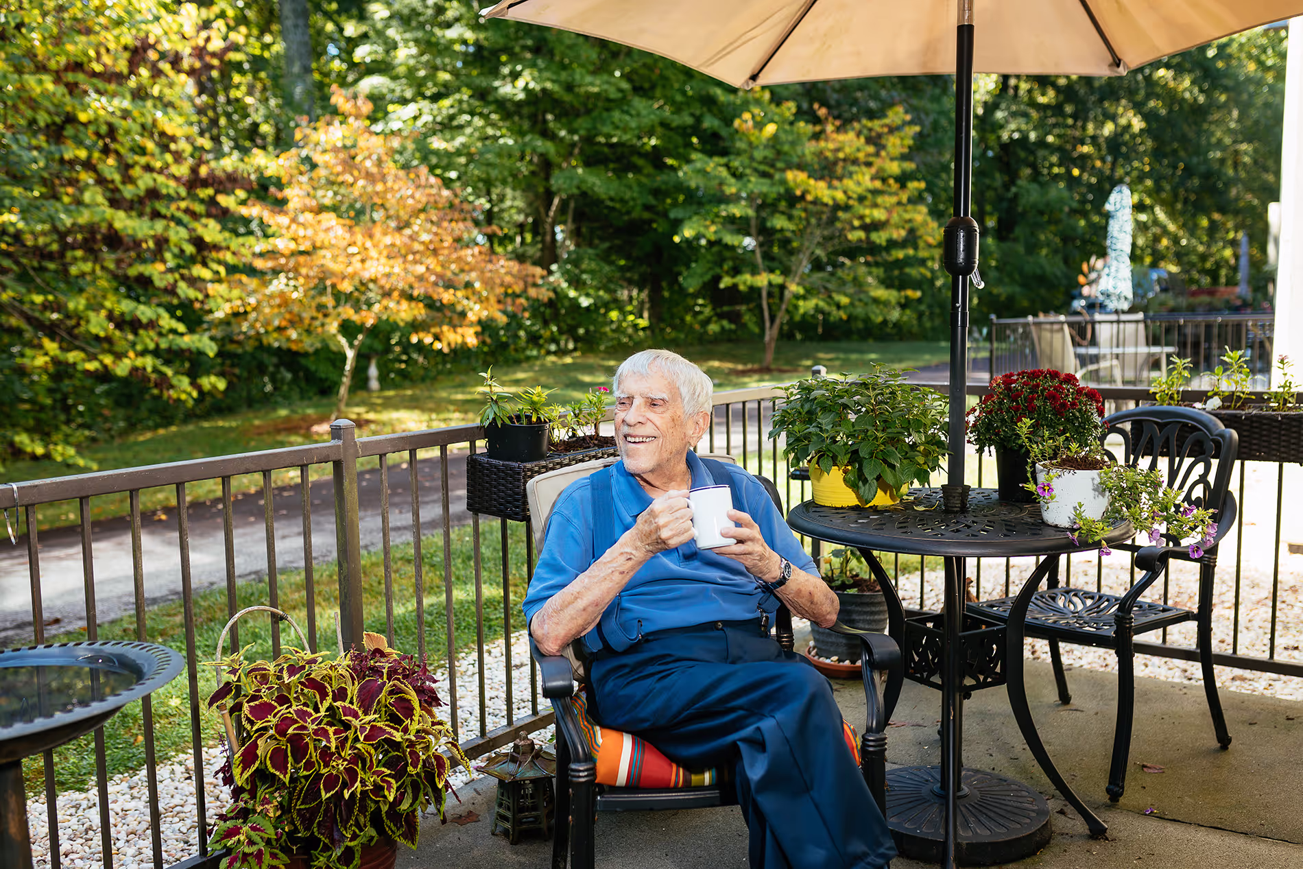 Elderly man sitting on a patio chair, smiling and holding a white mug under a large umbrella.