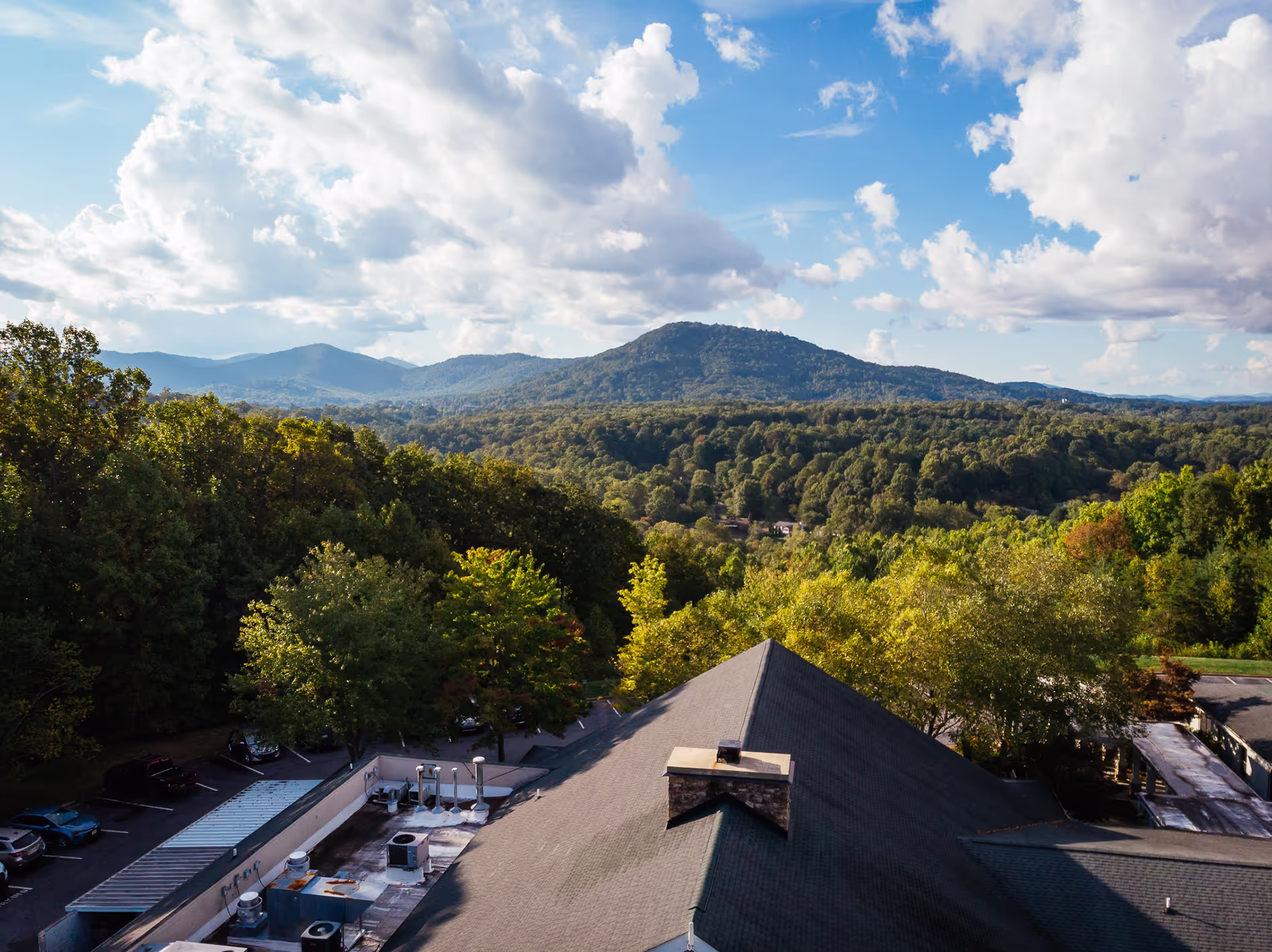 View of a mountain landscape with rooftops and trees in the foreground under a cloudy blue sky.