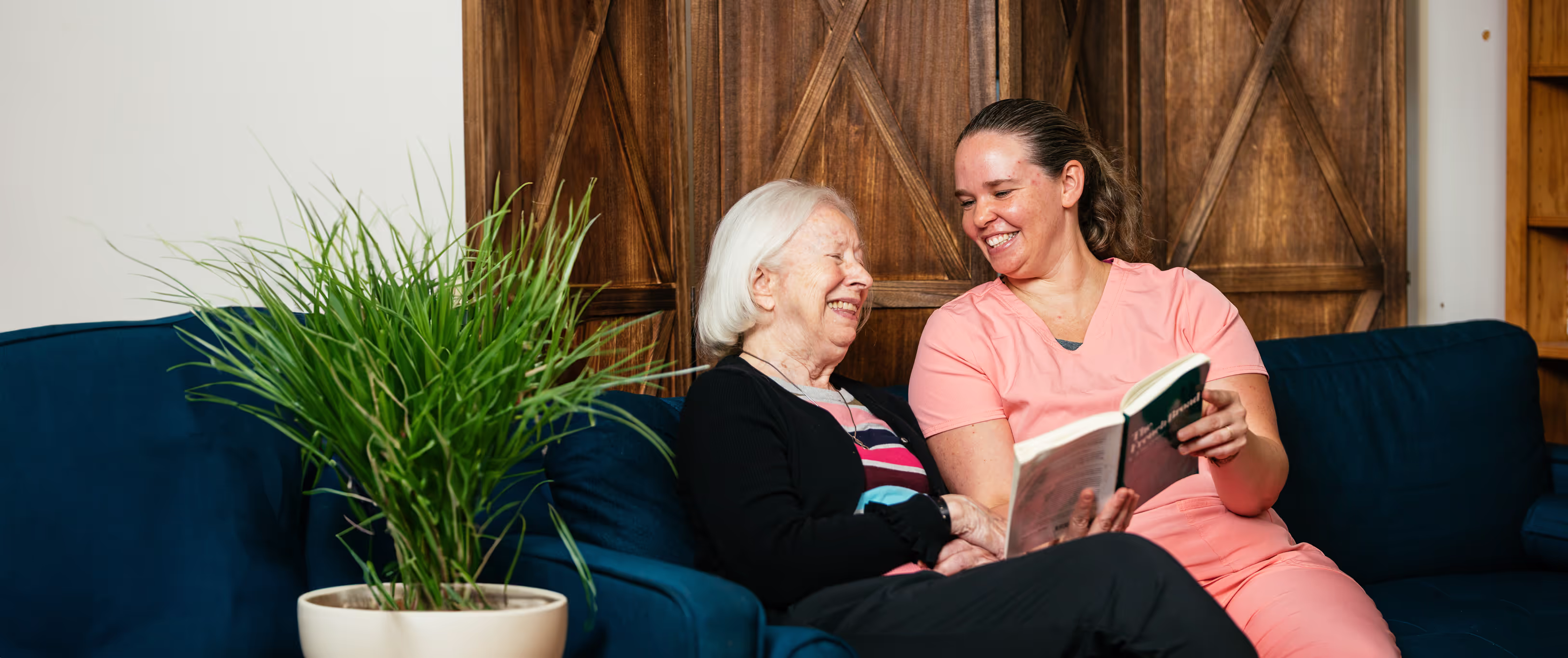 A young woman in scrubs reads a book to an elderly woman on a sofa.