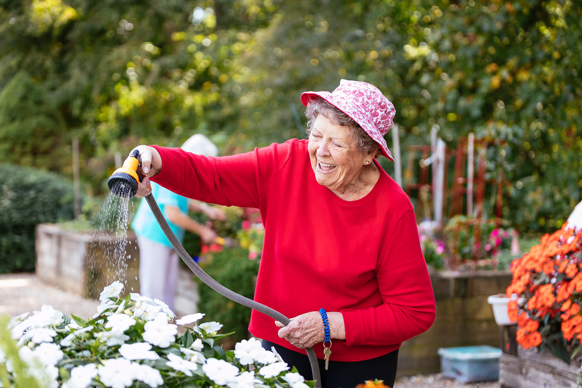 Elderly woman watering white flowers in a garden, wearing a red sweater and pink hat.