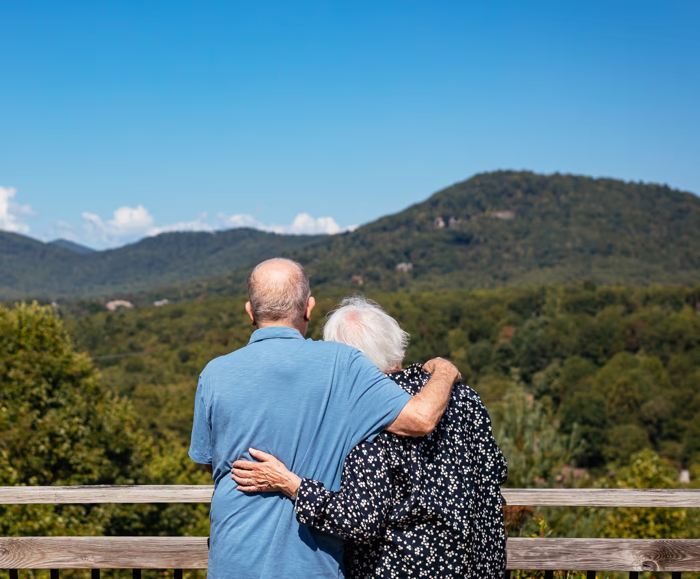 An elderly couple stands on a balcony, hugging while enjoying a mountain view.