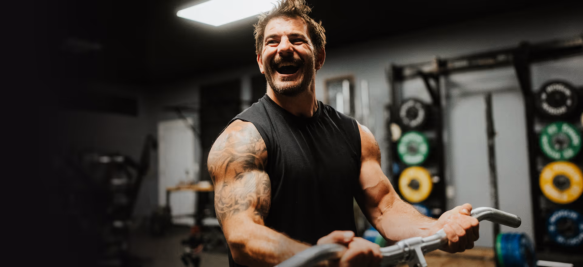 Smiling muscular man with tattooed arm lifting a barbell in a gym.