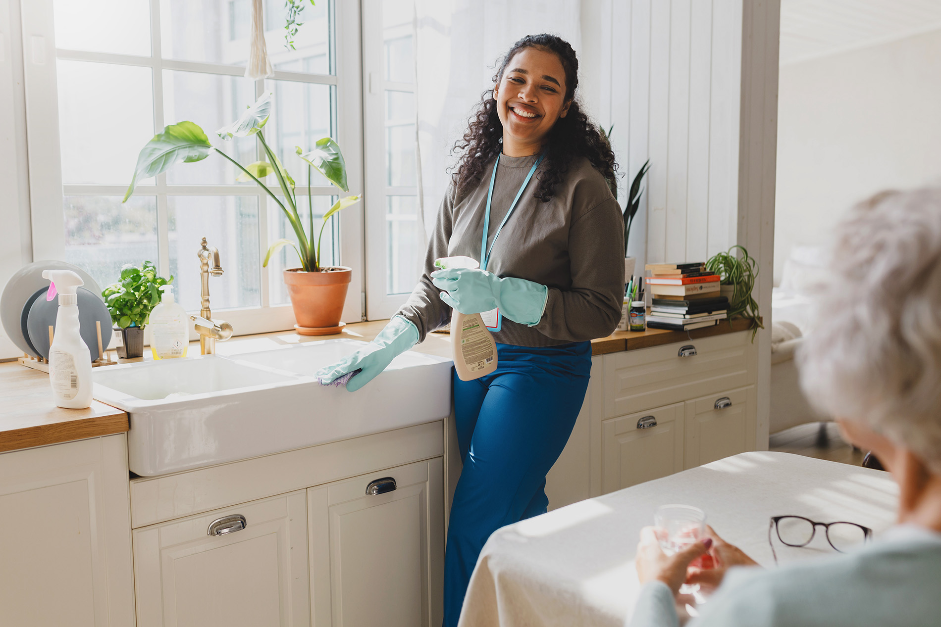 A woman wearing gloves smiles while cleaning a kitchen sink. An elderly person sits nearby.