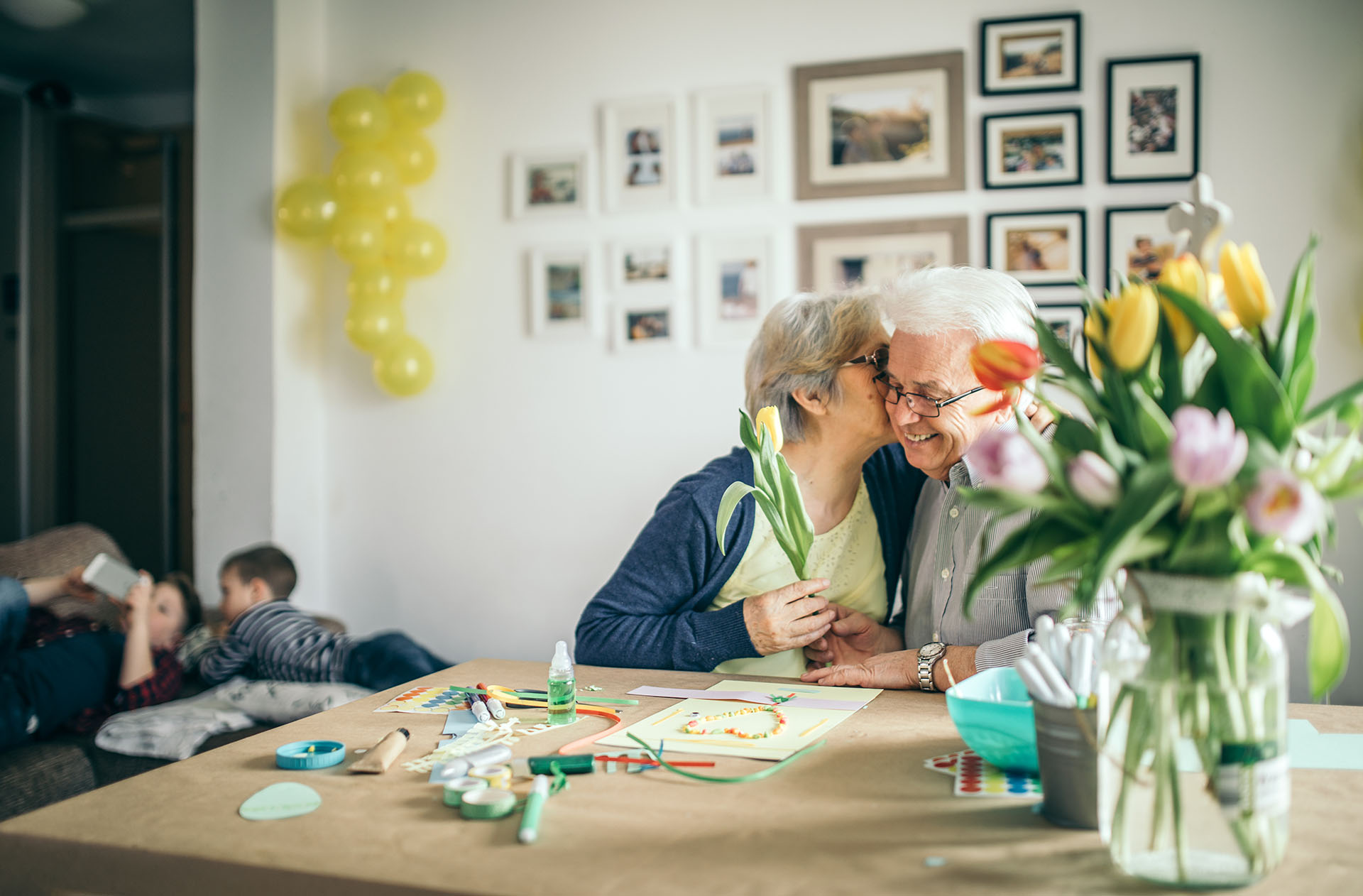 An elderly woman kisses an elderly man holding a tulip, with art supplies on the table.