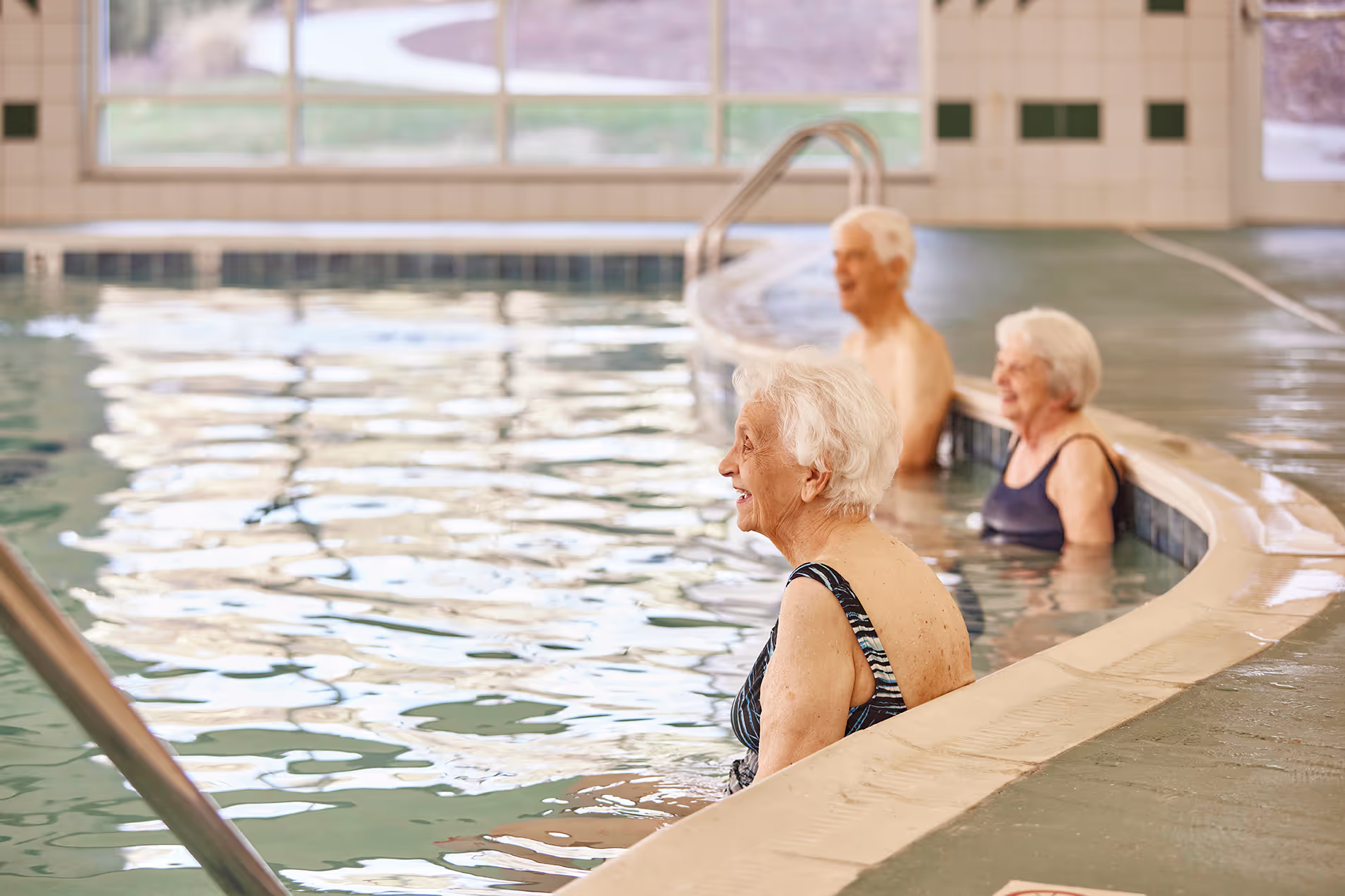 Three elderly people relax and chat in an indoor swimming pool near the edge.
