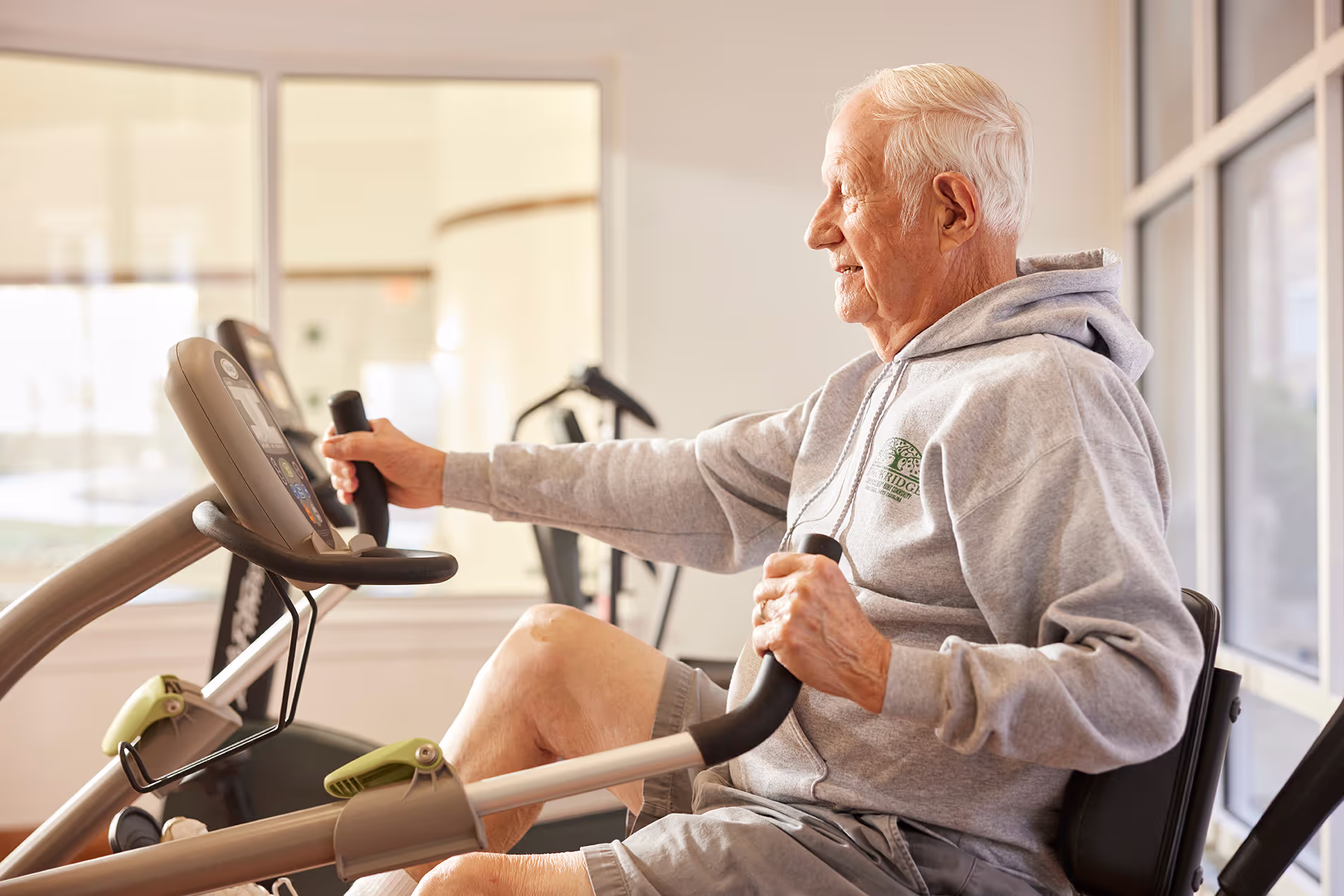 Older man wearing a hoodie exercises on a stationary bike indoors.