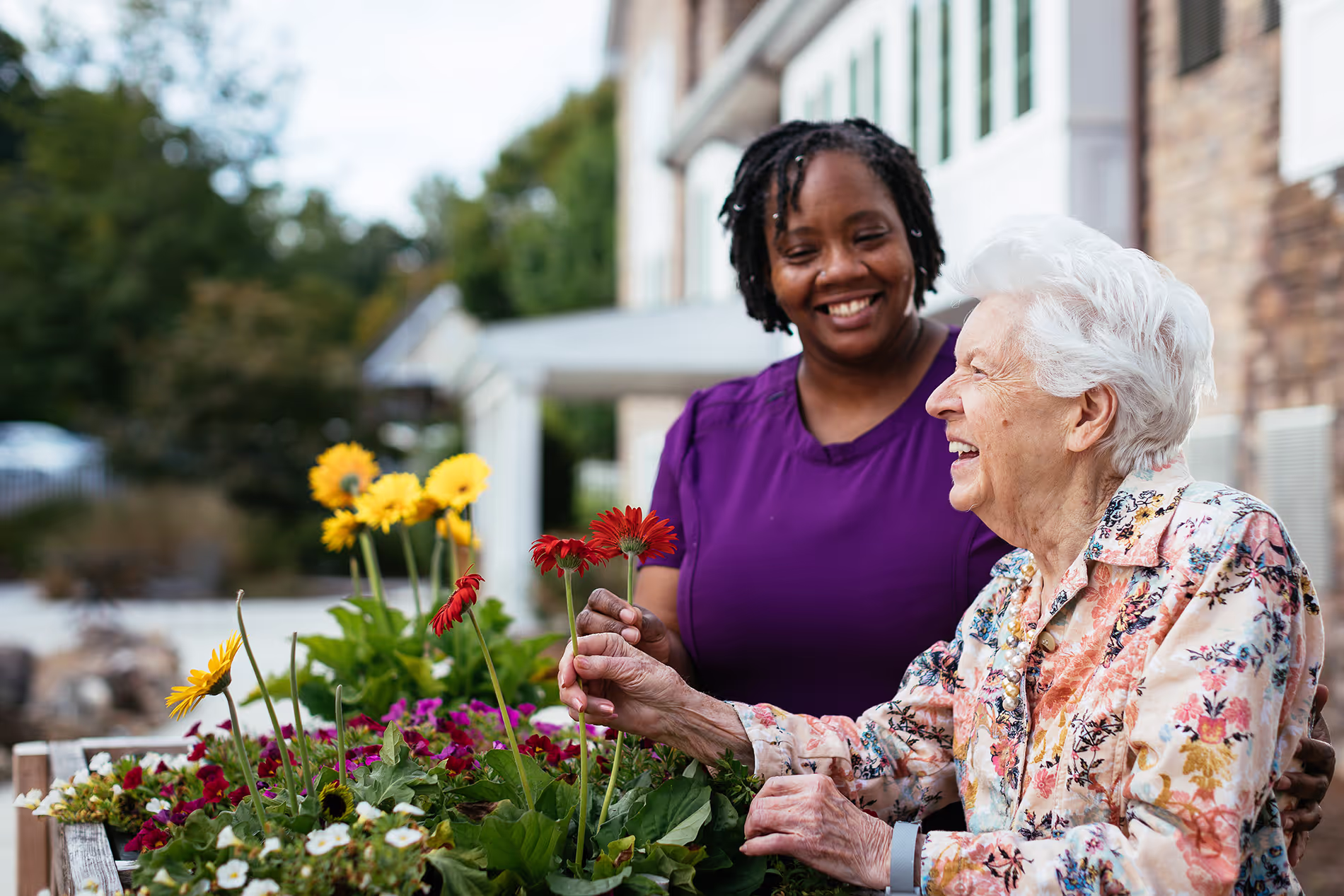 Two women smiling and planting flowers together in an outdoor garden.