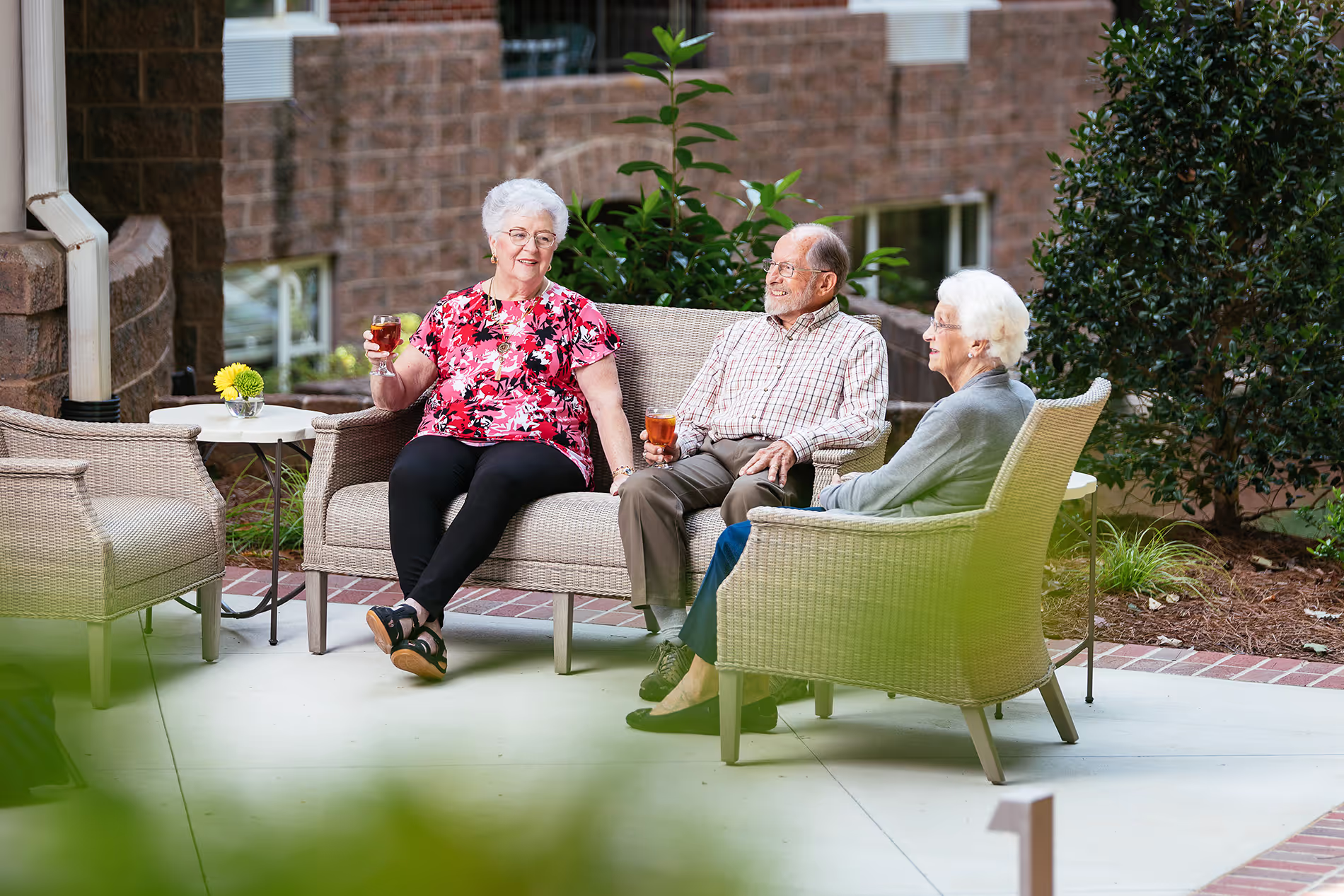 Three elderly people sit on patio chairs, enjoying drinks and chatting outside a building.