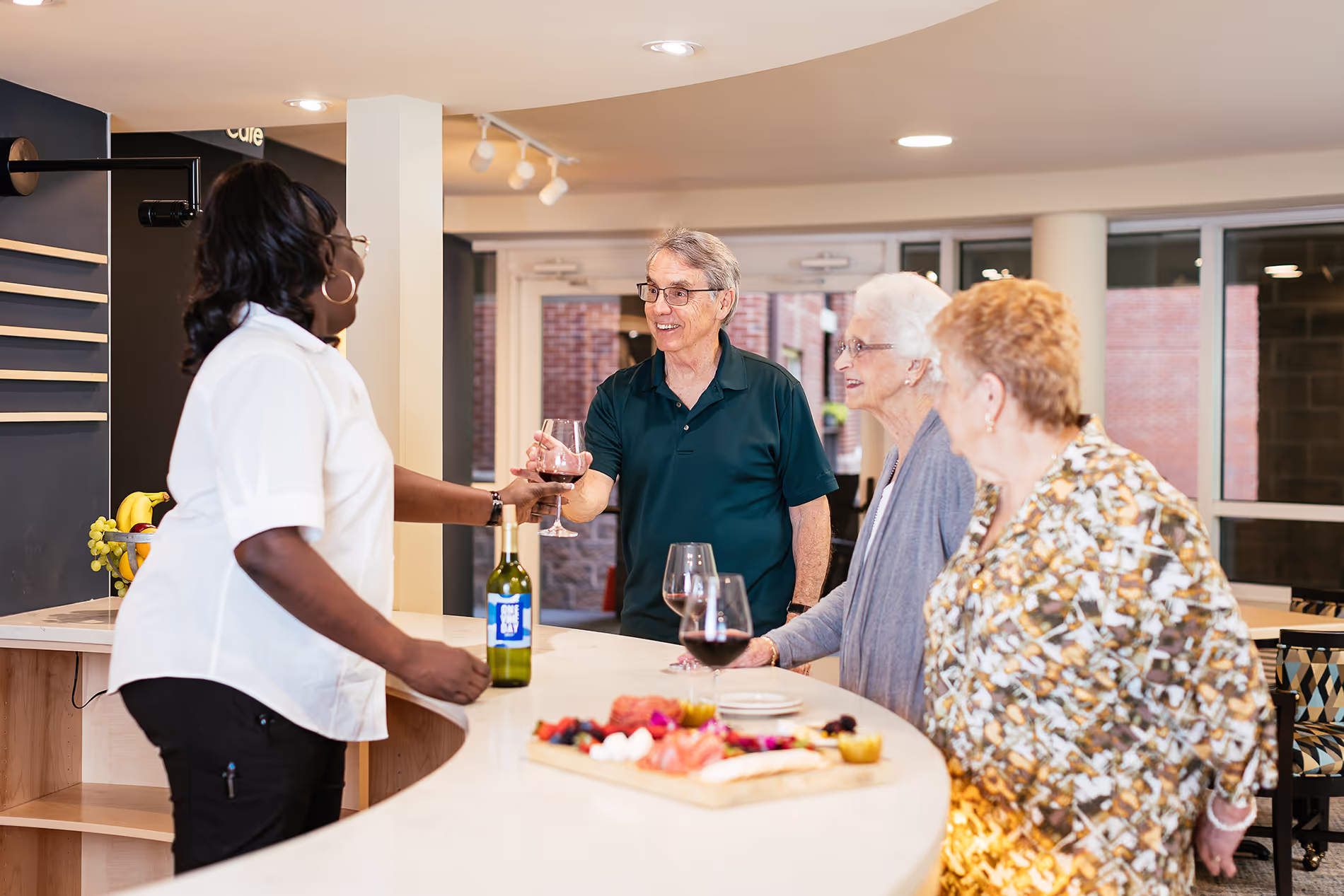A woman serves wine to three smiling older adults standing at a counter indoors.