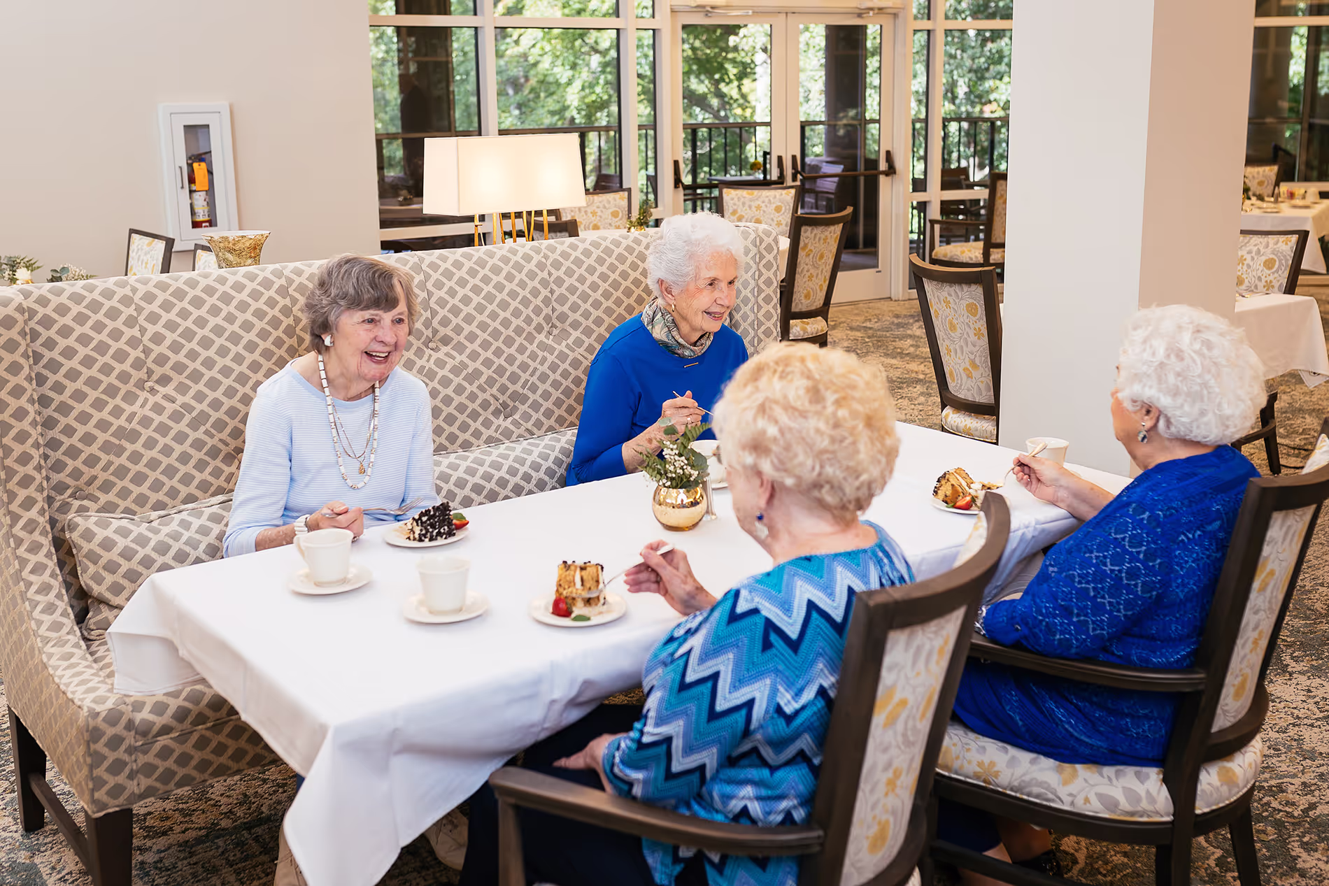 Four elderly women enjoy coffee and cake, sitting together at a dining table indoors.