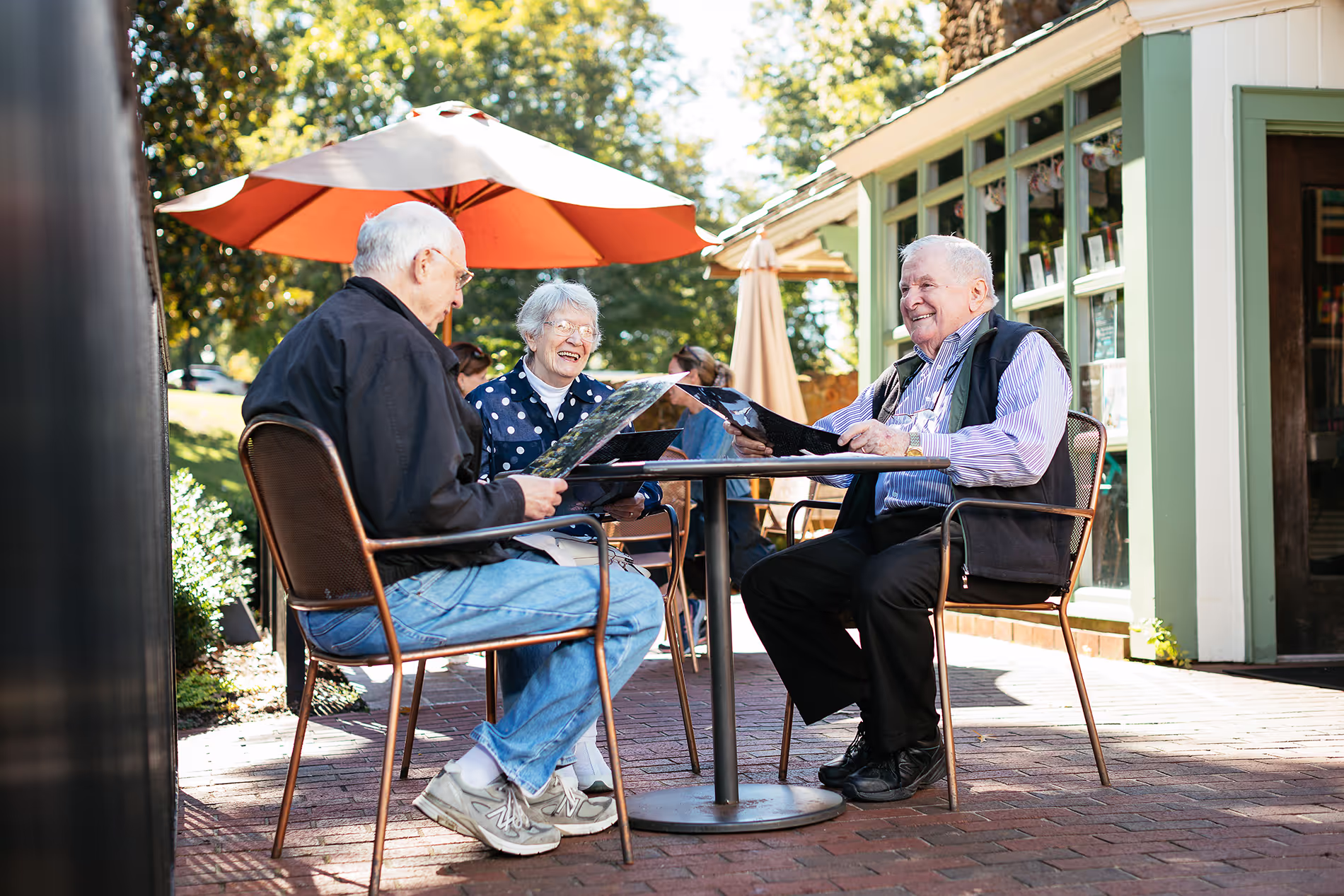 Three older adults sitting at an outdoor café table, smiling and reading menus together.