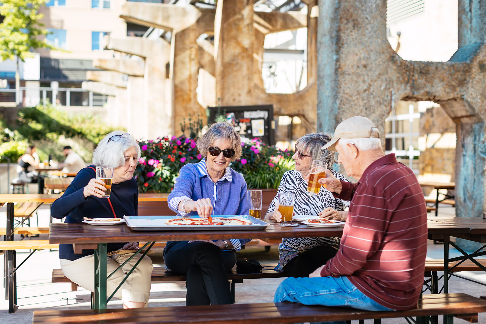 Four people sit at a picnic table, enjoying drinks and pizza outdoors.